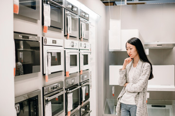 Half length portrait of thoughtful asian woman choosing new oven in furniture store