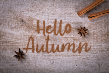 Hello, Autumn. The phrase is written on a wooden table with ground cinnamon among cinnamon sticks and badyan.  Top view, flat lay