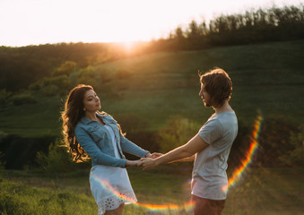 Fototapeta na wymiar Stylish young couple. Film photo at sunset and with a sunlight. A guy and a girl are holding hands against the background of the mountains. The girl has a white short dress and a jeans jacket