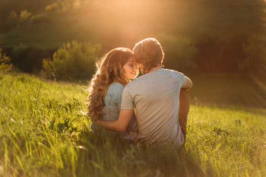 Stylish young couple sitting on a hill and admiring the sunset. A film photo with a light and a sunlight, a foreshortening from the back. Enamored youth in the second before the kiss