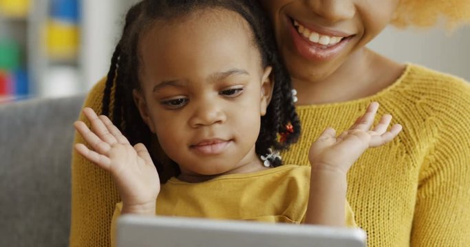 Close up of the little cute African American girl sitting on the knees of her mother, watching something on the screen of tablet and clapping hands. Indoors