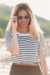 Happy blonde lady holds her glasses and laughting while walking on the sunny street. Outdoor portrait of glad fashionable woman wears shorts and t-shirt in summer warm day