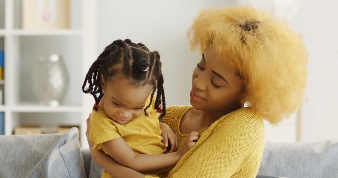 Portrait shot of the African American young mother and cute little daughter sitting on the gray sofa in the cozy living room, kissing and hugging. Inside