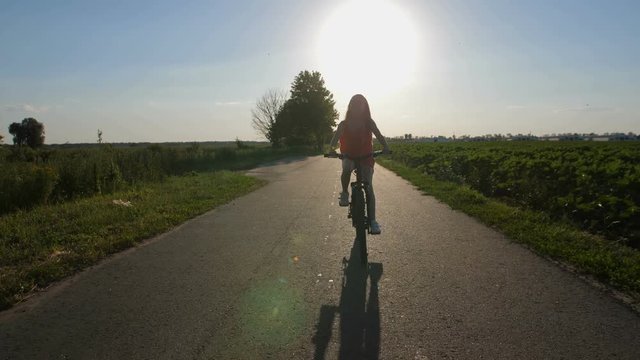 Attractive teen girl enjoying summer holidays and riding bicycle on countryside road through green fields. Positive and peaceful teenager in nature. Sun flare during sunset. Steadicam shot
