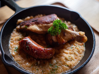 Sizzling hot, rich, slow-cooked Trio Cassoulet of duck confit, pork belly and smoked sausage on a bed of white beans in a black cast iron pan on a wooden table. Natural Light.