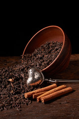 Dried black tea in clay bowl with sticks of cinnamon and a tea strainer isolated on black background