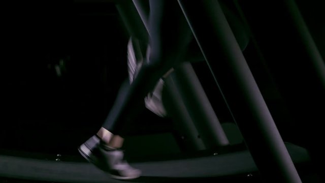 Woman s legs in sneakers walking then running on a treadmill in a dark gym. Side view Handheld real time medium shot