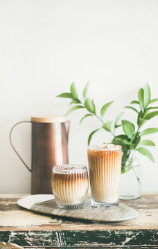 Iced coffee in tall glasses with milk on board, white wall, green plant branches and jug at background, copy space. Summer refreshing beverage ice coffee drink concept