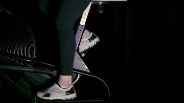 Woman s legs in white sneakers using a bicycle machine in a dark gym. Side view Concept of fitness. Left to right pan real time medium shot