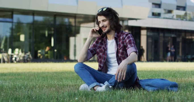 Caucasian young male student in the jeans and motley shirt sitting on the grass near college building and speaking on the phone. Outdoors.