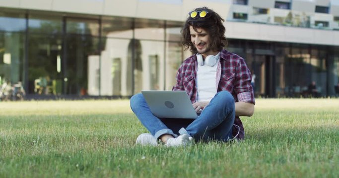 Caucasian young man in the motley shirt typing on his laptop computer and sitting on the grass near his college. Outside.