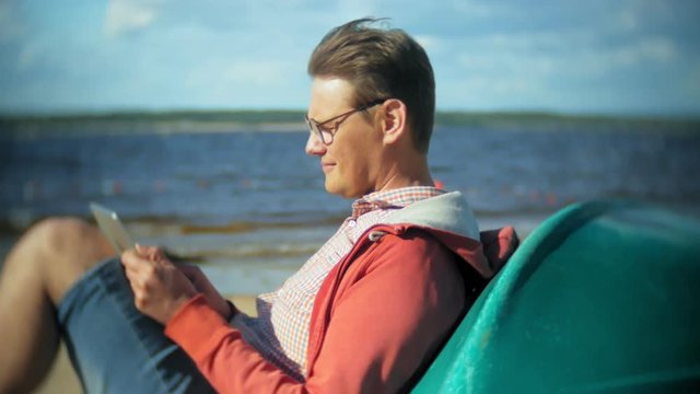 Old man sits on the beach in headphones near the boat and uses a tablet
