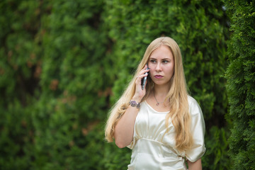 Young pretty blonde girl in a dress is talking on the phone between thuja on the street in summer.