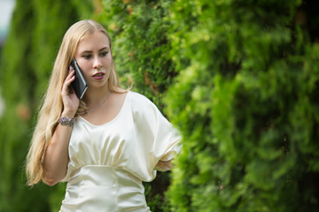 Young pretty blonde girl in a dress is talking on the phone between thuja on the street in summer.