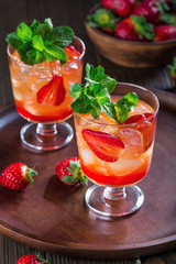 Red cocktail with ice, mint leaves and strawberry on wooden background.