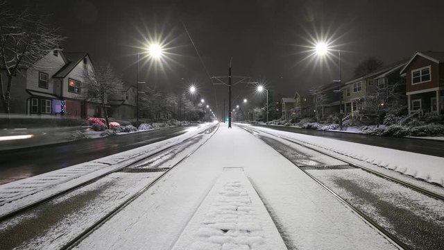 Light rail trains pass by either side of camera at night with light dusting of snow on ground and traffic moving on wet street.