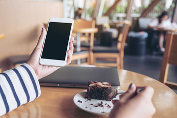 Mockup image of a woman holding white mobile phone with blank black desktop screen while enjoy eating brownie cake in cafe