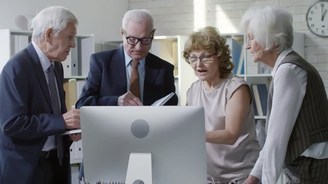 Medium shot of team of senior businessmen and businesswomen in formalwear looking at computer screen and discussing work