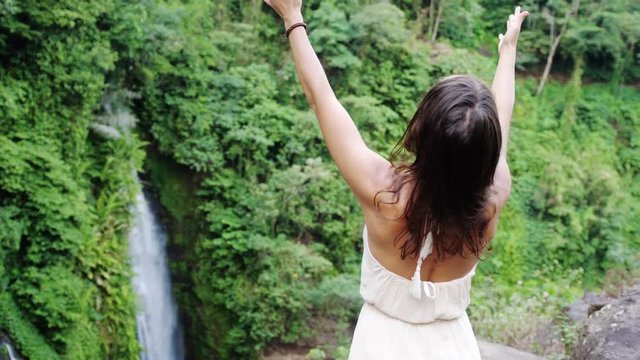 Young brunette woman in long dress rises hands at waterfall in jungle Koh Samui. Thailand. slow motion. 3840x2160