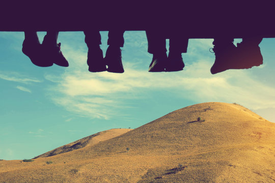 Underside view of dangling Four people legs with wearing trekking shoes sitting on the edge of wooden board-walk against a background of mountains and blue sky