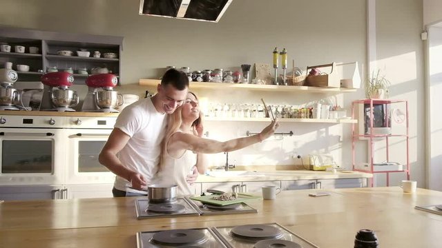 A nice tall european guy in a white shirt is trying a dish and hugging his wife in a white dress while she is taking pictures with her phone