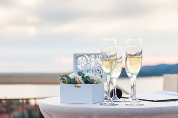 Wineglasses of ice champagne on table for wedding reception after wedding ceremony by the sea and sunset. Box with flowers with rings.