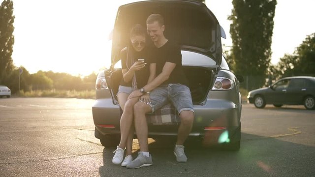 Happy coule sitting in the open car trunk. Pretty girl with deadlocks and sunglasses scrolling her smartphone while her boyfriend caress her legs