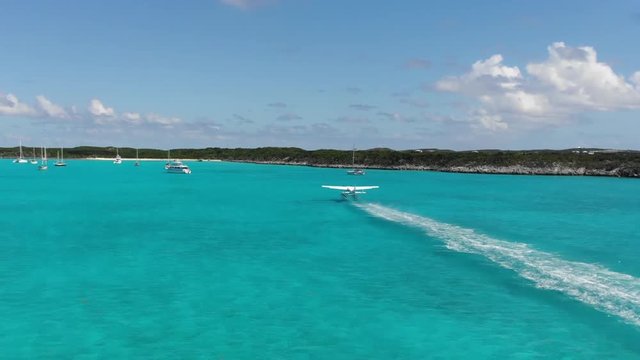 Water plane takes off from harbor surrounded by sailboats, fishing boats, and yachts. Located in a nest of islands in the Bahamas. Aerial drone follows plane at takeoff.