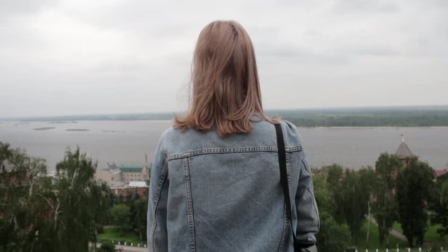Back view of young teenage woman in jeans jacket with long hair standing and looking at view of wide Volga river in Nizhny Novgorod