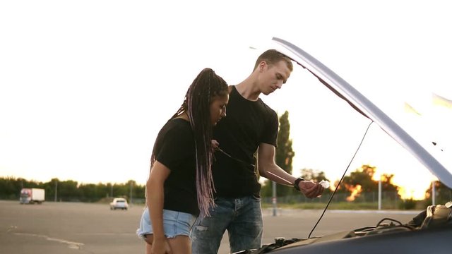 Side view of a young couple stopped while their road trip on the parking zone. Young man in black T shirt checks the oil level, his girlfriend with dreadlocks standing near the open car hood. Parking