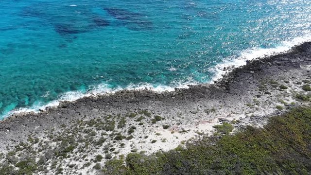 Aerial drone view of waves crashing into rocky island shore before panning up the coast line. Forested island and white sandy beach are visible from overhead drone shot.