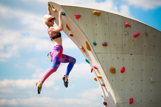 Photo from side of sports blonde in leggings hanging on wall for climbing against blue sky