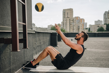 side view of handsome sportsman doing sit ups with medicine ball on yoga mat on roof