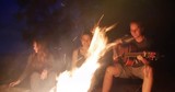 Young man singing songs with guitar and two girls sitting by the campfire in forest