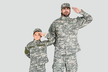 portrait of confident soldier and son in military uniforms saluting isolated on grey