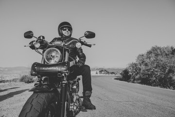 Black and white portrait of man with black helmet, jacket and sunglasses standing on a classic American motorcycle.