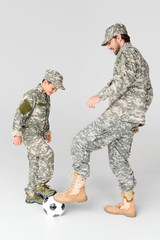side view of father and son in military uniforms playing soccer on grey background