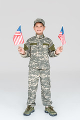 smiling child in military uniform with american flagpoles in hands looking at camera on grey...