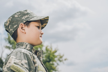 side view of little boy in camouflage clothing with cloudy sky on backdrop