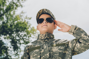 portrait of little boy in camouflage clothing and sunglasses saluting with cloudy sky on backdrop