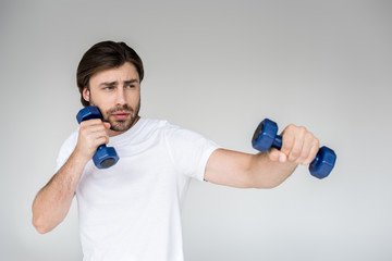 portrait of man in white shirt with blue dumbbells in hands exercising on grey backdrop