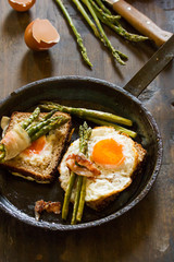 Delicious fried eggs on a toasted bread in a metal pan with grilled asparagus wrapped in fried bacon.