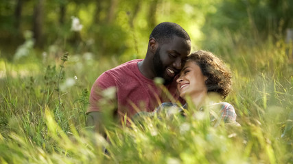 Multiracial couple laughing and chatting while lying in grass, outdoor date