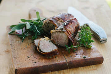 Delicious roasted pork fillet wrapped in herbs and ham with garlic, thyme, sage and rosemary in the background, on the wooden board.