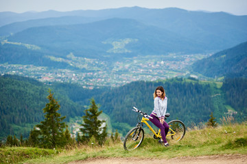 Obraz premium Young woman biker sitting on yellow bike near rural trail on the top of the mountain on cloudy evening. Mountains, forests and small city on the blurred background. Outdoor sport activity