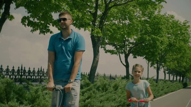 Modern active family with scateboard scooters resting in summer park. Joyful millennial father and lovely daughter riding kick scooters together while family spending weekend outdoors. Slow motion.