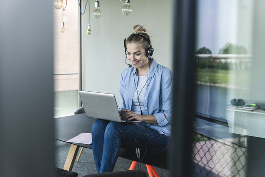Young businesswoman sitting at desk, using headset and laptop