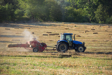Obraz premium Farmer baling hay in the field. Baling hay with a conventional hay baler in the field. Straw bale pressing machine