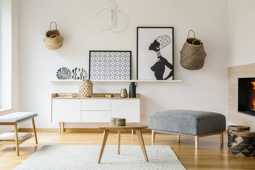 Posters and baskets on white wall in bright flat interior with wooden table next to pouf. Real photo