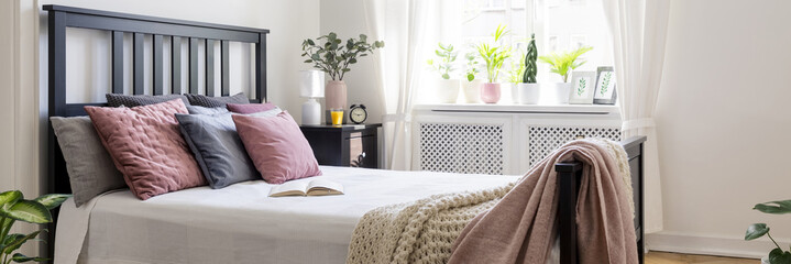 Real photo of bright bedroom interior with plants on windowsill, open book on king-size bed with pastel cushions and black bedhead and window with curtains